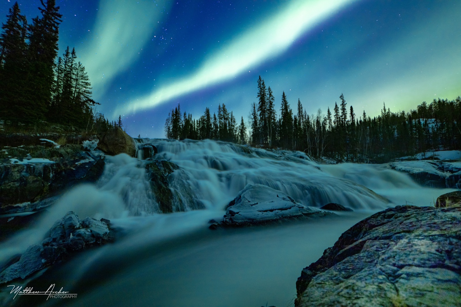 Green aurora arcing over a wide northern waterfall, the water smoothed by a long exposure with spruce forest on the far bank