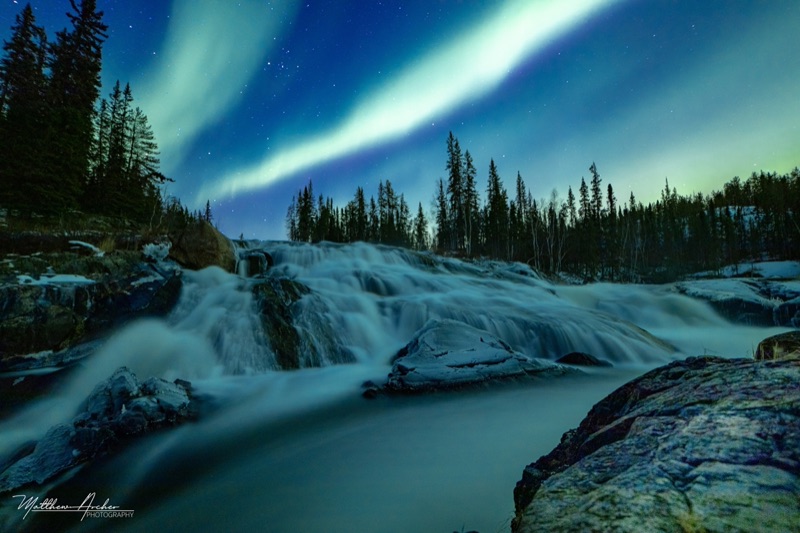 Green aurora arcing over a wide northern waterfall, the water smoothed by a long exposure with spruce forest on the far bank