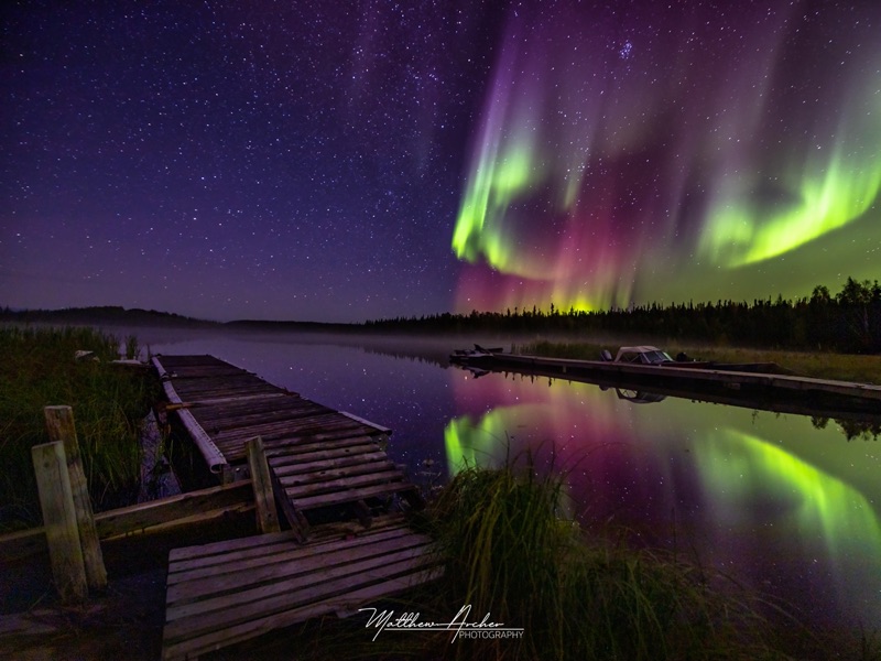 Pink and green aurora mirrored in a still lake beside an old wooden dock