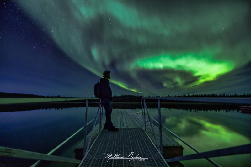 A figure standing at a dock railing looking out at a swirl of green aurora above a calm lake