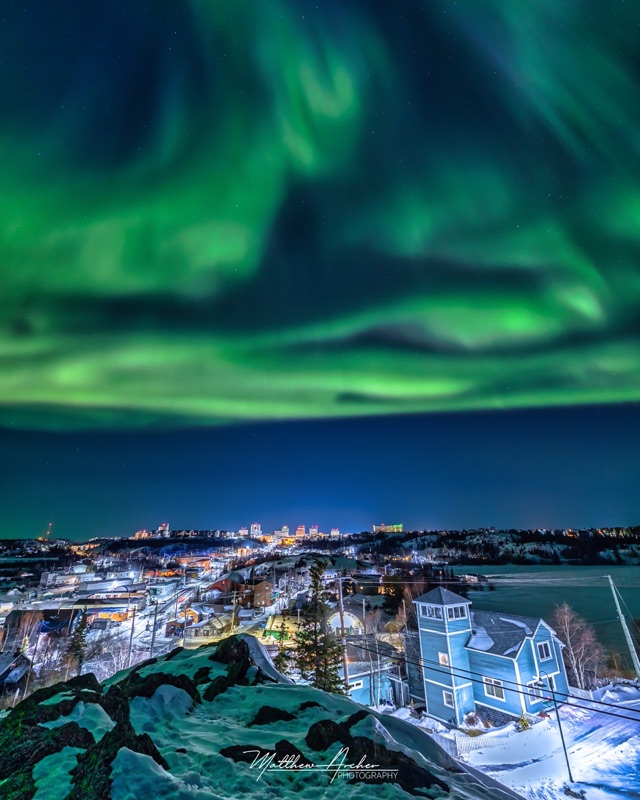 Aurora sweeping over Yellowknife at night, the lit downtown and houses below a snow-covered rock outcrop