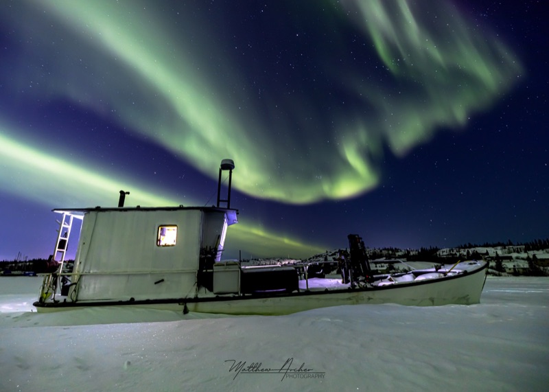 A derelict fishing boat locked in lake ice, one cabin window lit, green aurora overhead