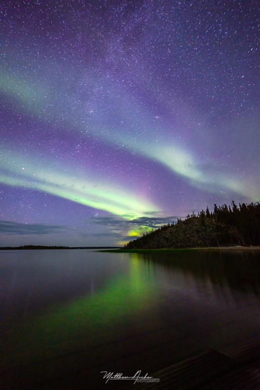 Green and violet aurora with the Milky Way above a calm lake, the colour running down the water toward the shore