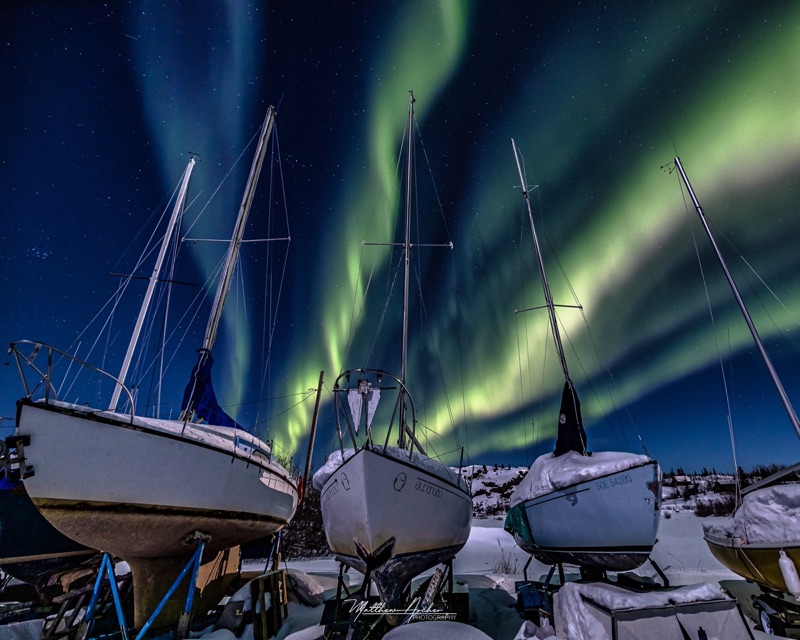 Sailboats in winter storage under snow, masts rising into a sky streaked with green aurora