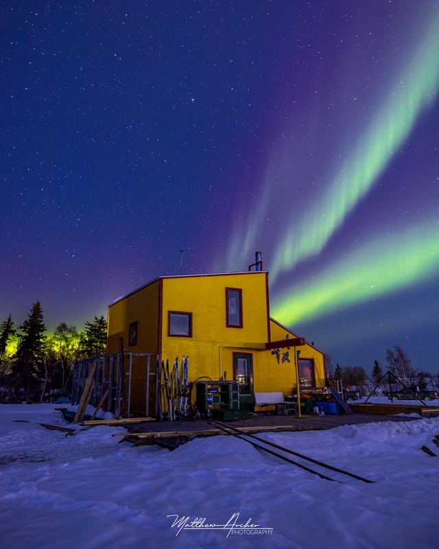A yellow two-storey houseboat sitting on the ice with green aurora arcing overhead