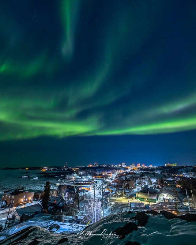 Green aurora over Yellowknife seen from a rock outcrop, the lit downtown spread out below
