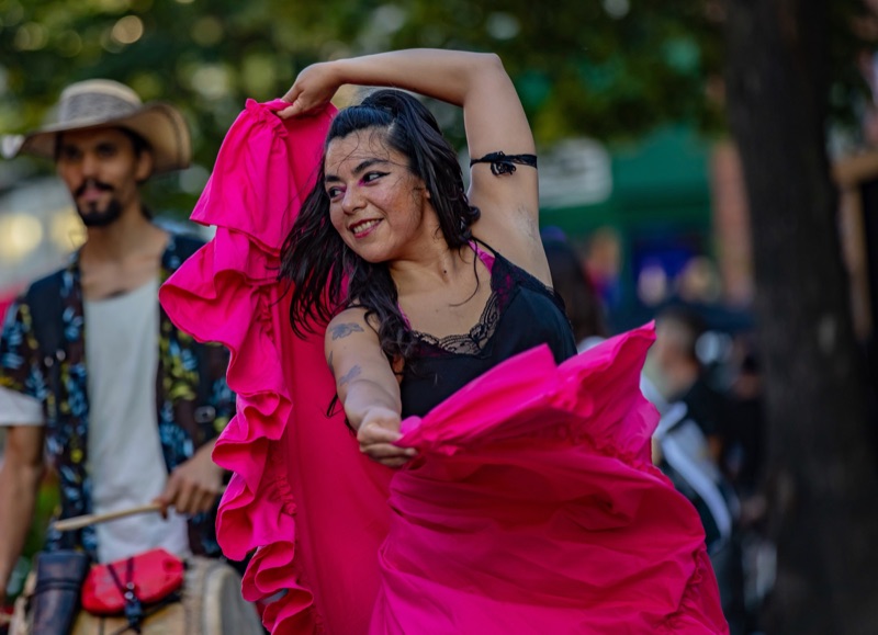 A dancer in a vivid pink ruffled dress mid-turn at an outdoor festival, one arm raised, a drummer playing behind her