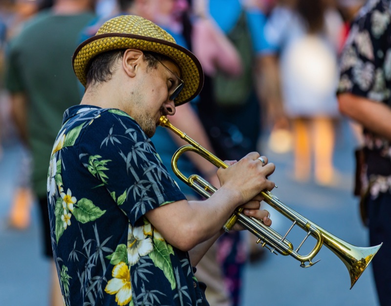 A trumpet player in a straw hat and floral shirt performing on a busy street, the crowd blurred behind him