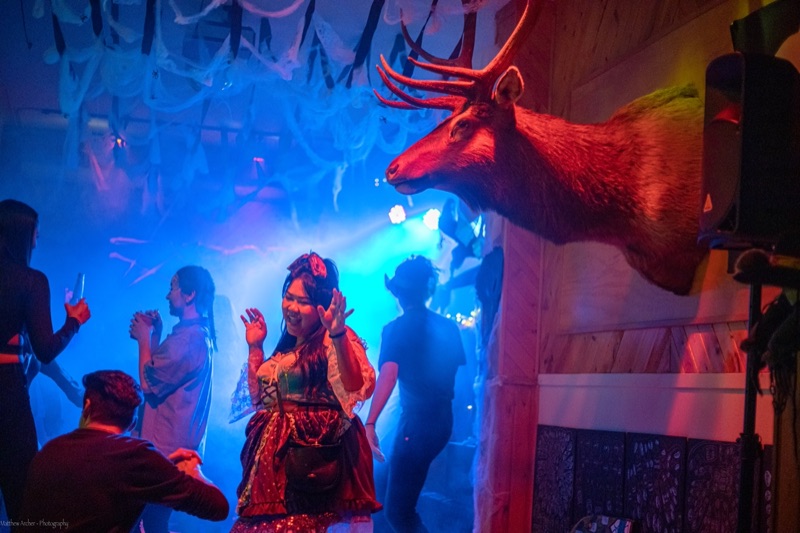 Dancers under blue light at a Halloween party, cobwebs strung from the ceiling and a mounted elk head lit red in the foreground