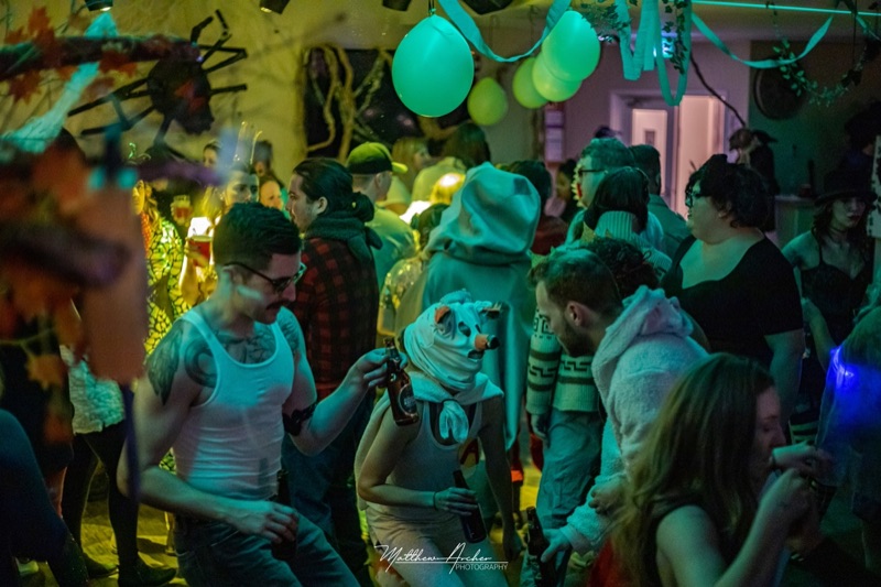 A crowded Halloween house party under green light, guests in costume with drinks in hand and balloons overhead