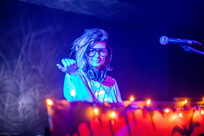 A DJ in oversized glasses reaching out from behind the decks, lit red and blue with string lights along the booth