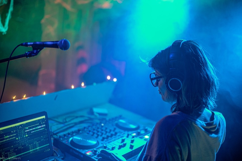 A DJ in headphones working a controller beside a laptop, backlit in blue and green haze