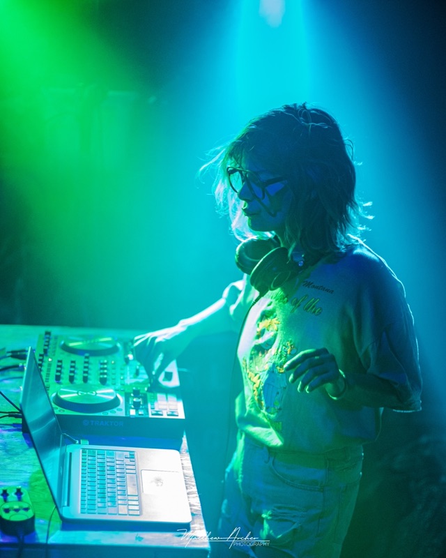 A DJ in glasses working a controller and laptop under teal light