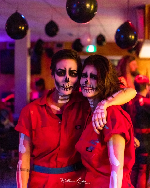Two people in skeleton face paint and red coveralls at a Halloween party, black balloons overhead and red light across the room