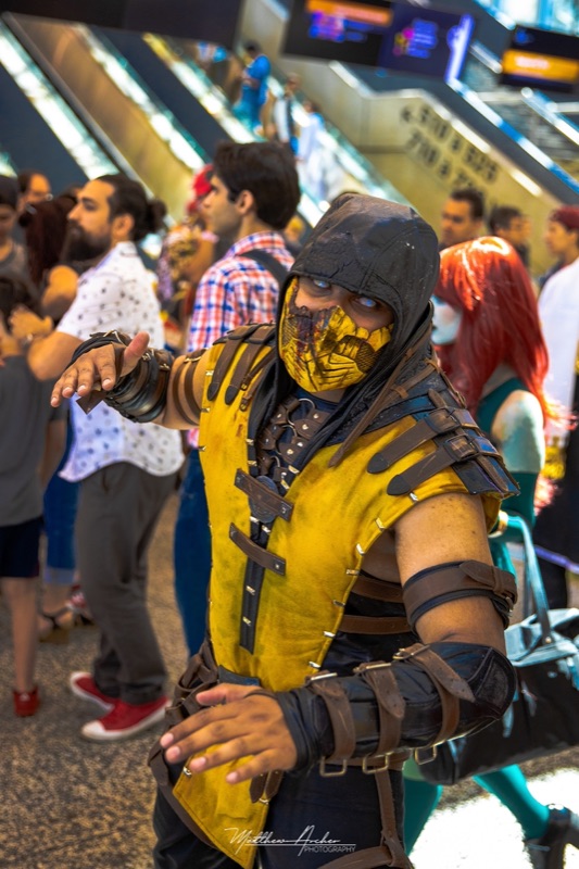 A cosplayer in full Scorpion costume and mask posing in a crowded convention hall