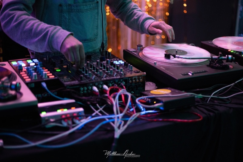 A DJ's hands over a mixer and turntables, cables running across the booth with warm lights behind