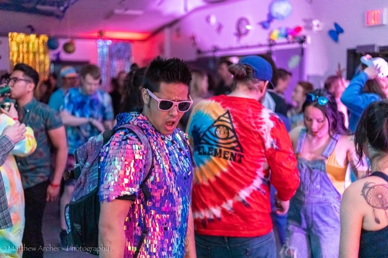 A partygoer in a sequinned jacket and white sunglasses grinning on a busy dance floor