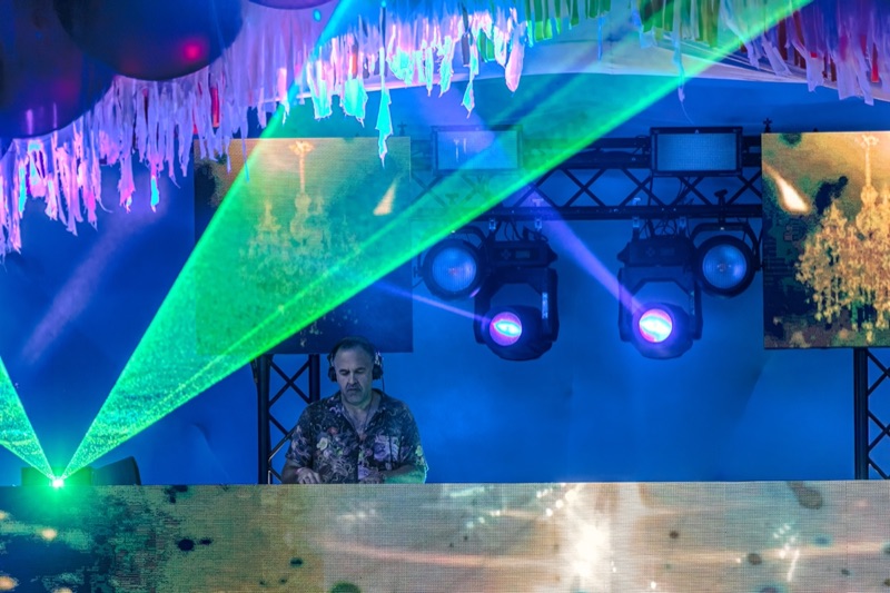 A DJ framed by green lasers and hanging streamers on a blue-lit festival stage