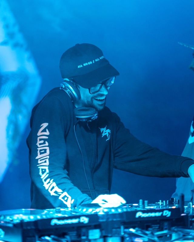A DJ in a cap and headphones grinning over the decks, bathed in deep blue light