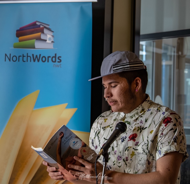 A reader in a flat cap and floral shirt reading from a book at the microphone beside a NorthWords banner