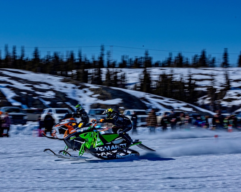 A snowmobile racer cornering hard across a frozen lake, spectators watching from the snowbank behind