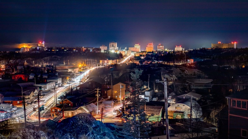Yellowknife at night seen from a hillside, headlights drawn into long light trails along the road and the downtown towers lit against a deep blue sky