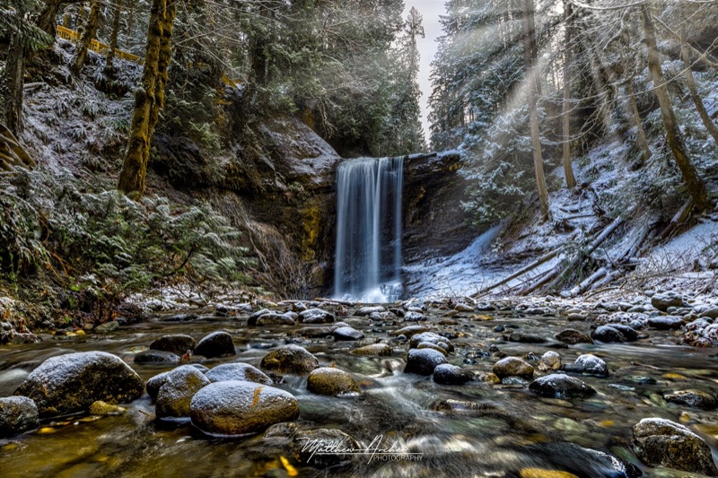 Ammonite Falls dropping into a snow-dusted forest canyon, sunbeams slanting through the trees above a boulder-strewn creek
