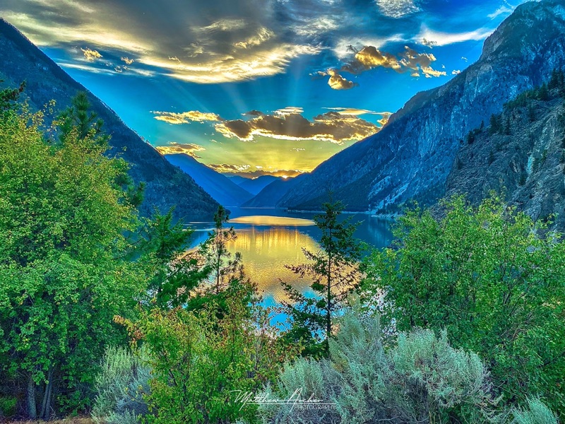 Sunbeams breaking through cloud over Duffey Lake at sunset, the mountains on either side mirrored in still water below