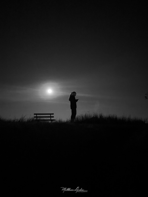 Black and white photograph of a person standing in silhouette on a grassy ridge beside an empty bench, the moon low and bright behind thin cloud