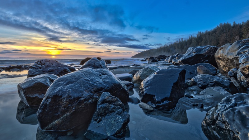 Wet boulders on Mystic Beach at sunset, a forested headland reaching into the water beneath a deep blue sky