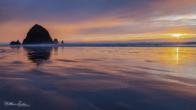 Haystack Rock silhouetted at sunset, wet sand reflecting the gold and purple sky