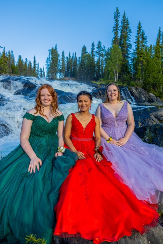 Three graduates in green, red and lilac gowns seated together on rock beside a rapids, spruce forest behind