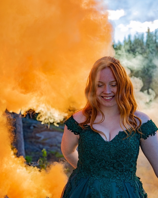 A graduate in a green gown laughing as orange smoke drifts across the frame behind her