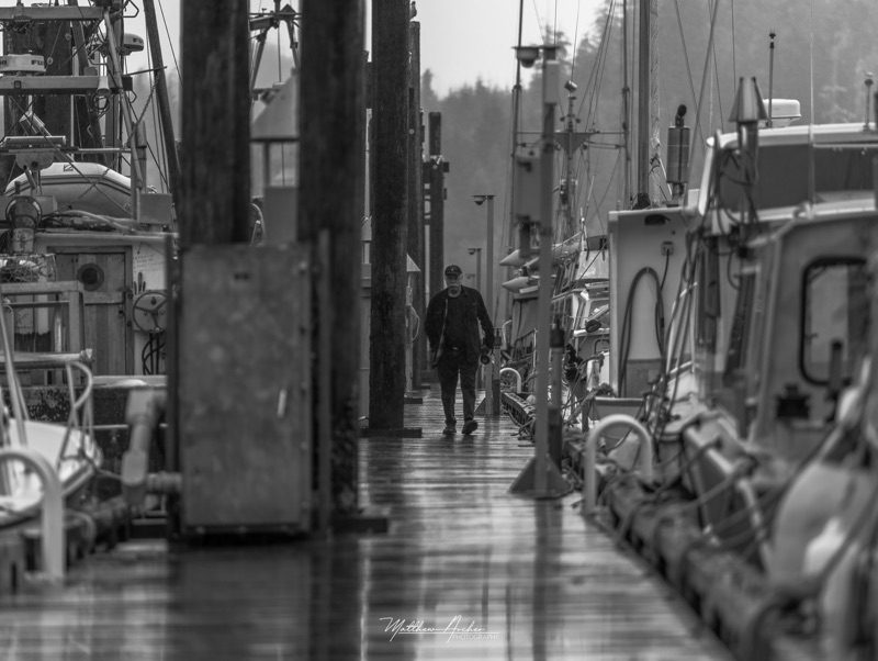 Black and white photograph of a lone figure walking a rain-slick fishing dock between moored boats