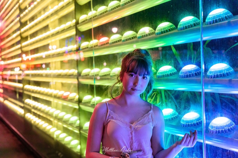 A young woman standing against a wall of coloured LED lights, the rainbow glow washing across her