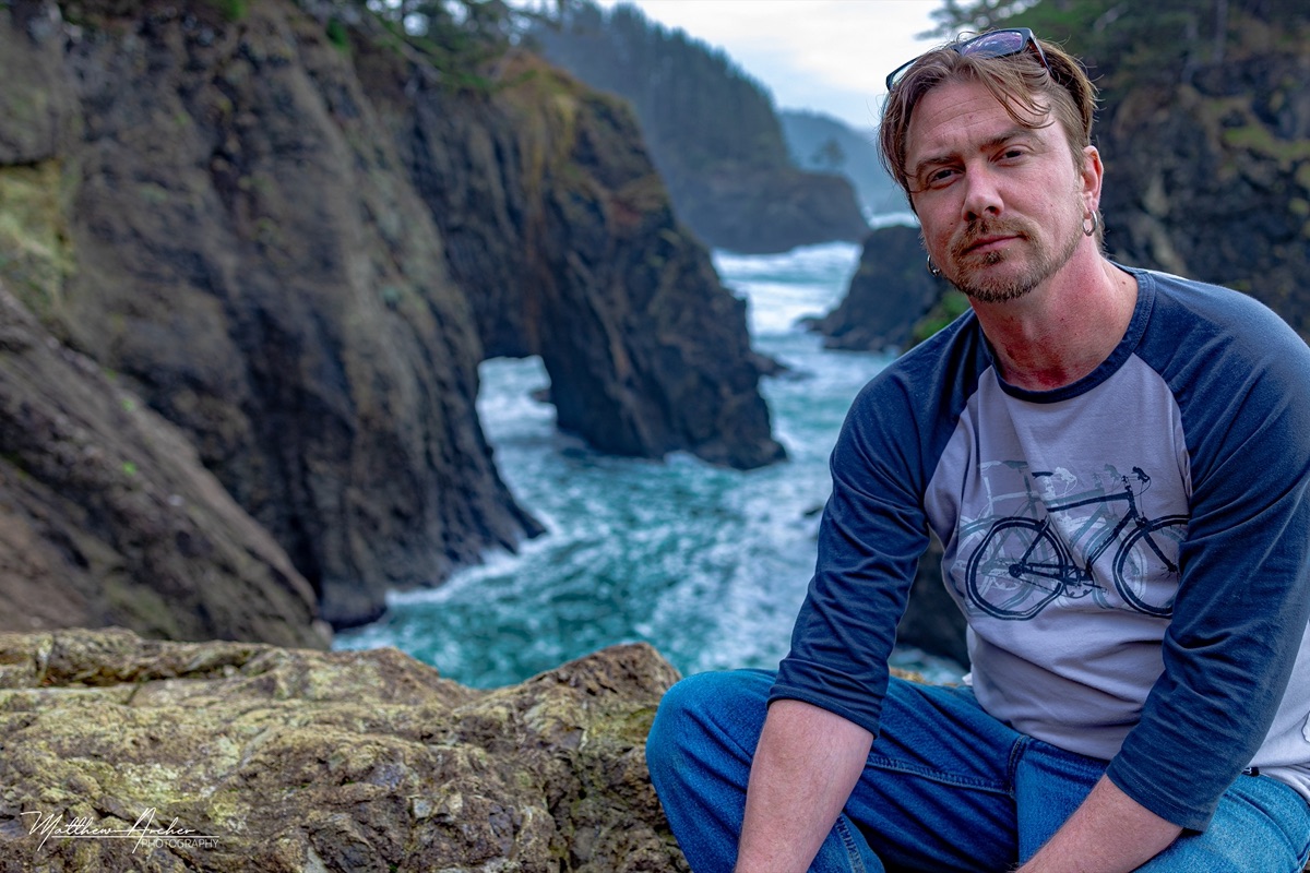 Matthew Archer sitting on a clifftop above a rocky coastline, surf breaking in the inlet below