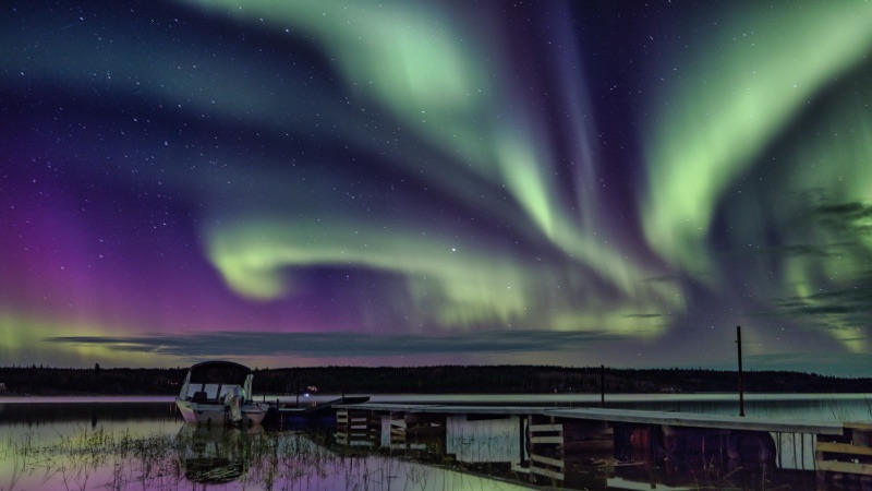Green and violet aurora wheeling above a quiet lake dock — "Nàaka over the Dock", a fine art print by Matthew Archer