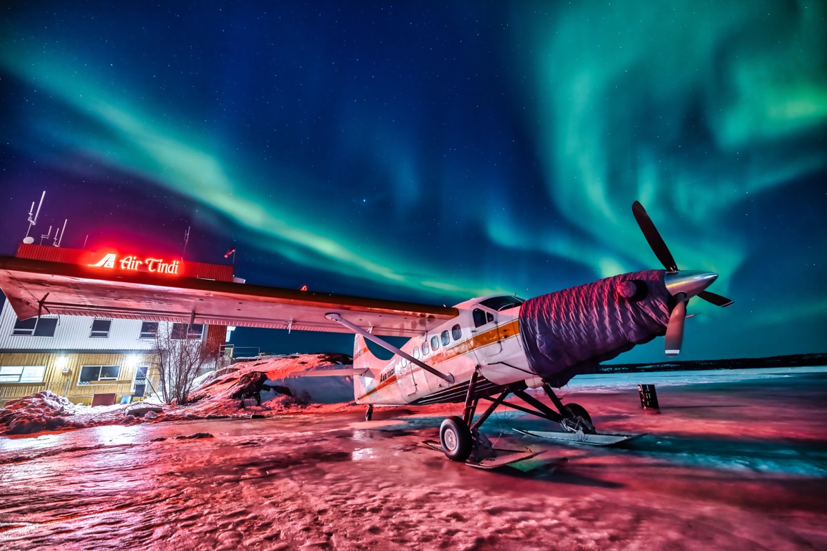 A ski-equipped bush plane under its engine blanket outside the Air Tindi hangar, aurora running the whole sky — "Blanket and Borealis", a fine art print by Matthew Archer