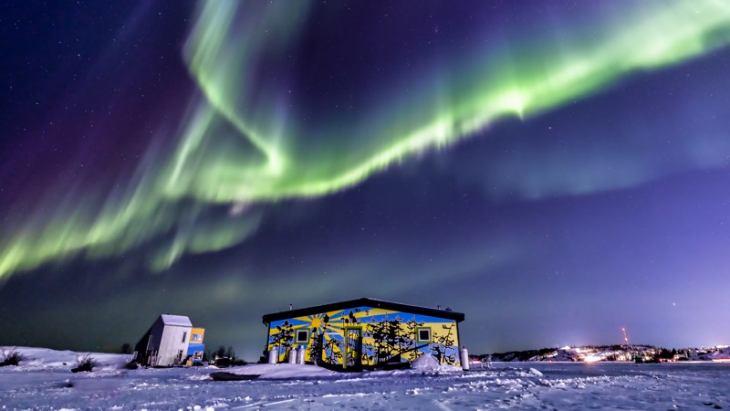 Aurora splitting into ribbons above a painted building on the snow, town lights low on the horizon — "Fractured Sky", a fine art print by Matthew Archer